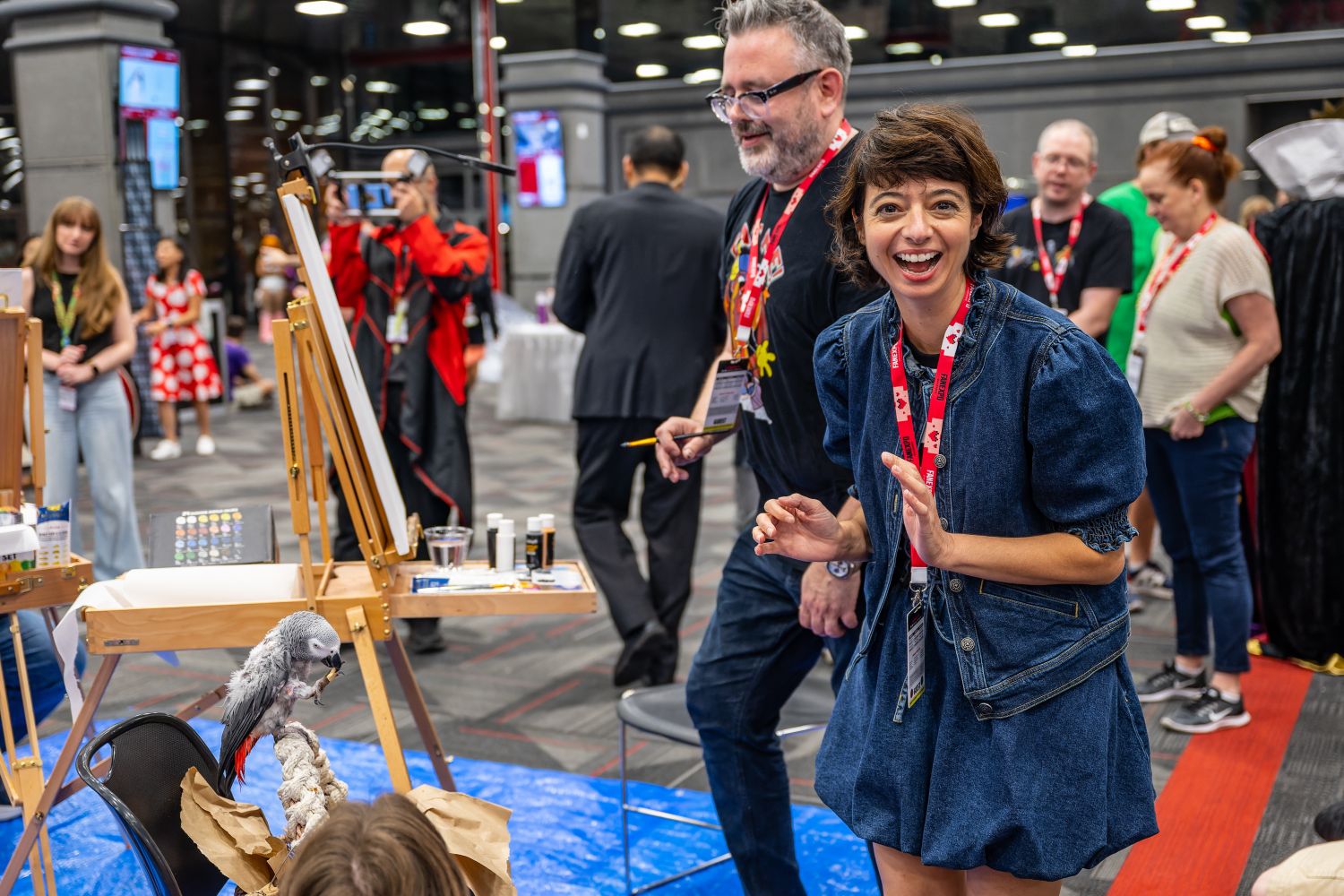 A woman in a denim dress smiles brightly toward the camera while standing near easels at the Comics Xplosion event. Behind her, another artist prepares to paint, and a parrot perches on a rope in the foreground.