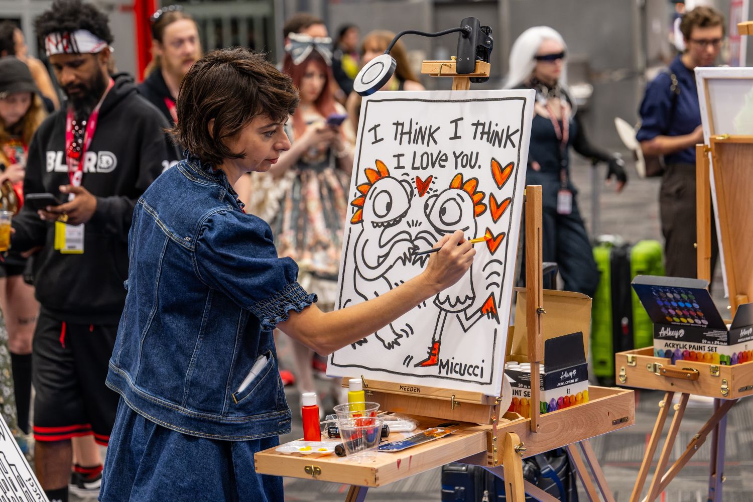 A woman in a denim dress paints a playful cartoon of two birds with the words “I THINK I THINK I LOVE YOU” written above them. Bright orange hearts decorate the piece as attendees look on.