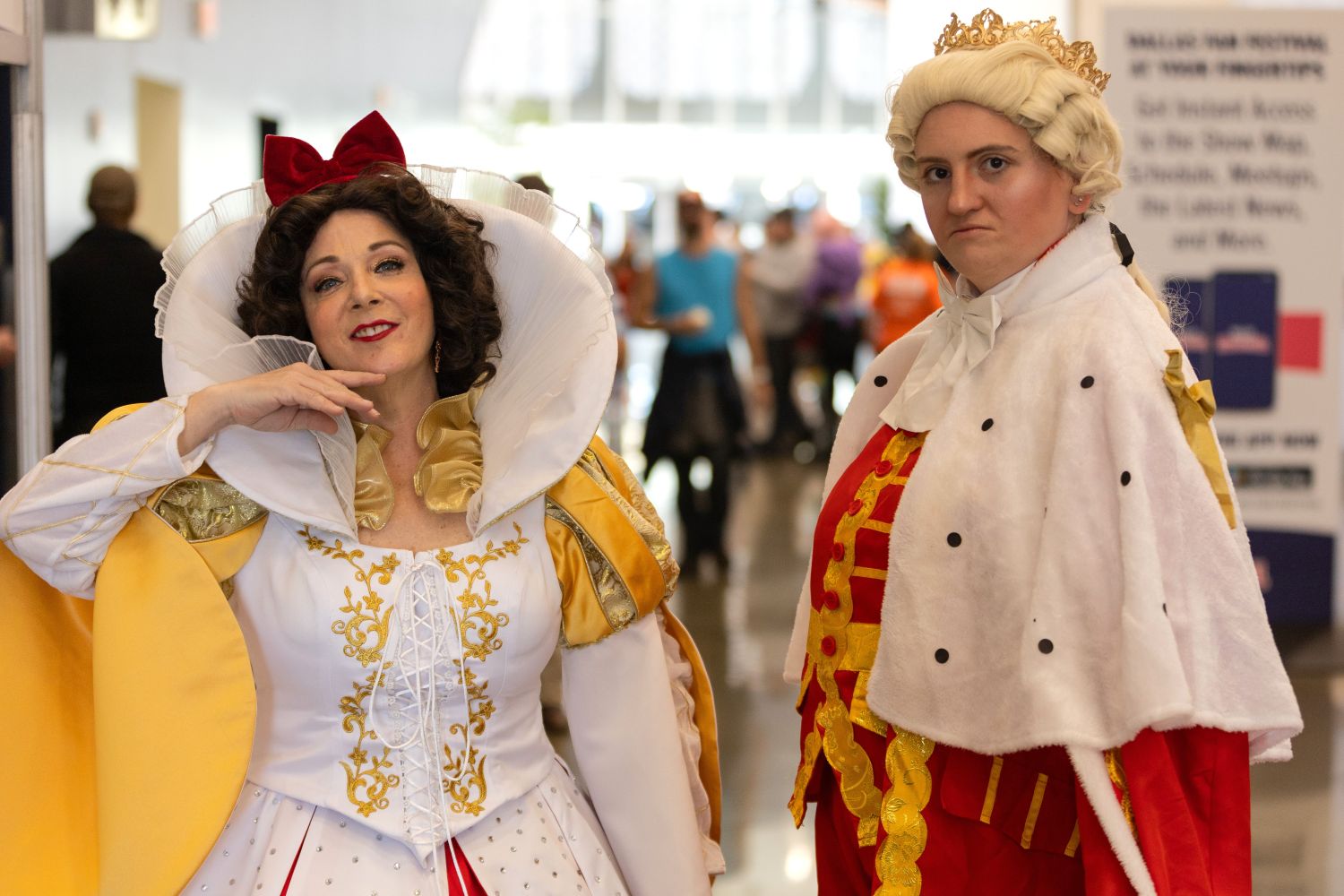 Two cosplayers pose in elaborate costumes at Dallas FAN FESTIVAL. One is dressed as the Evil Queen from Snow White with a dramatic gold and white gown, while the other wears a regal King George III outfit with a crown and red robe.