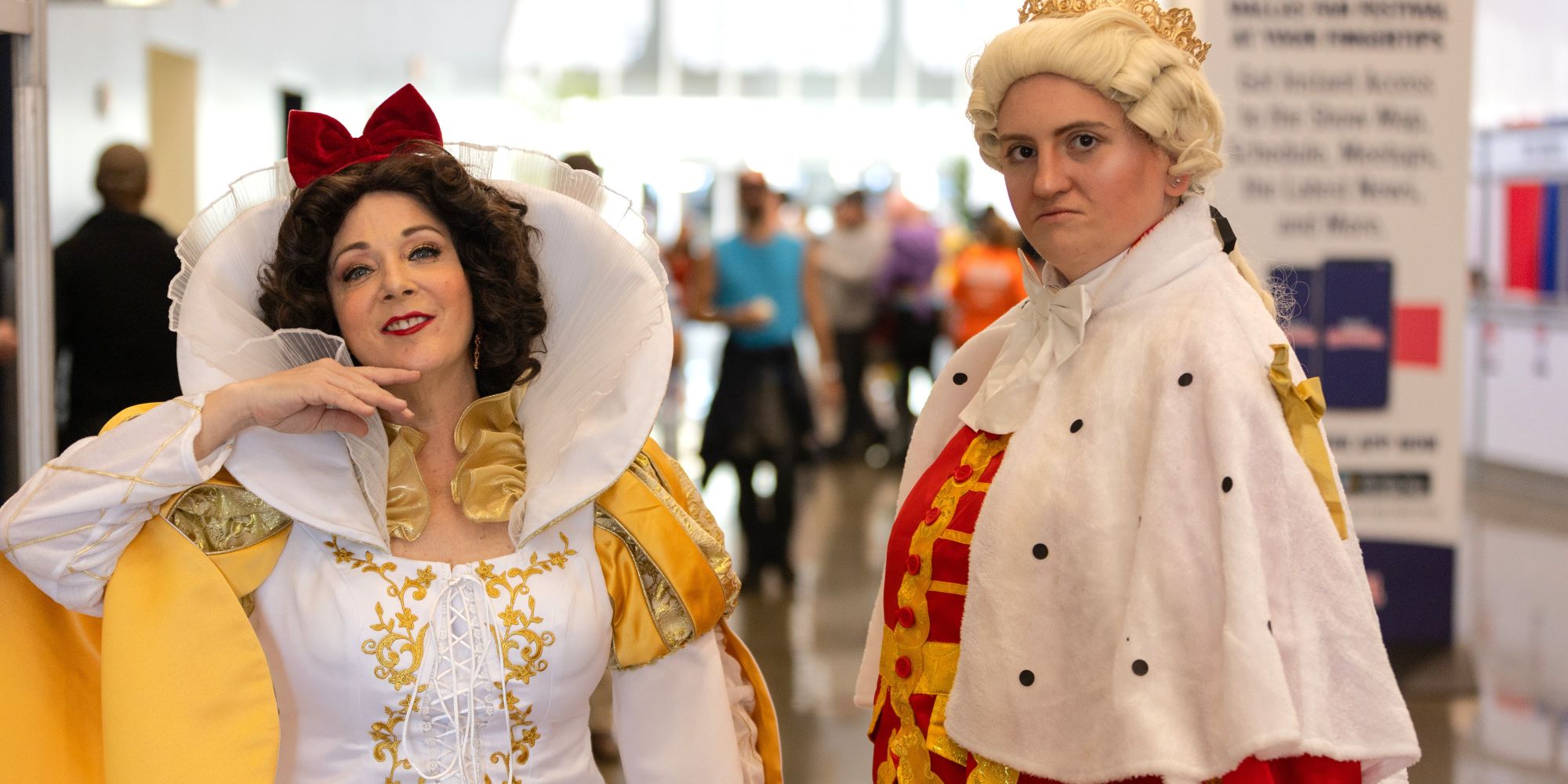 Two cosplayers pose at Dallas FAN FESTIVAL. One is dressed as the Evil Queen in a dramatic white and gold gown with a large red bow, while the other wears a regal King George III costume with a crown and red robe.