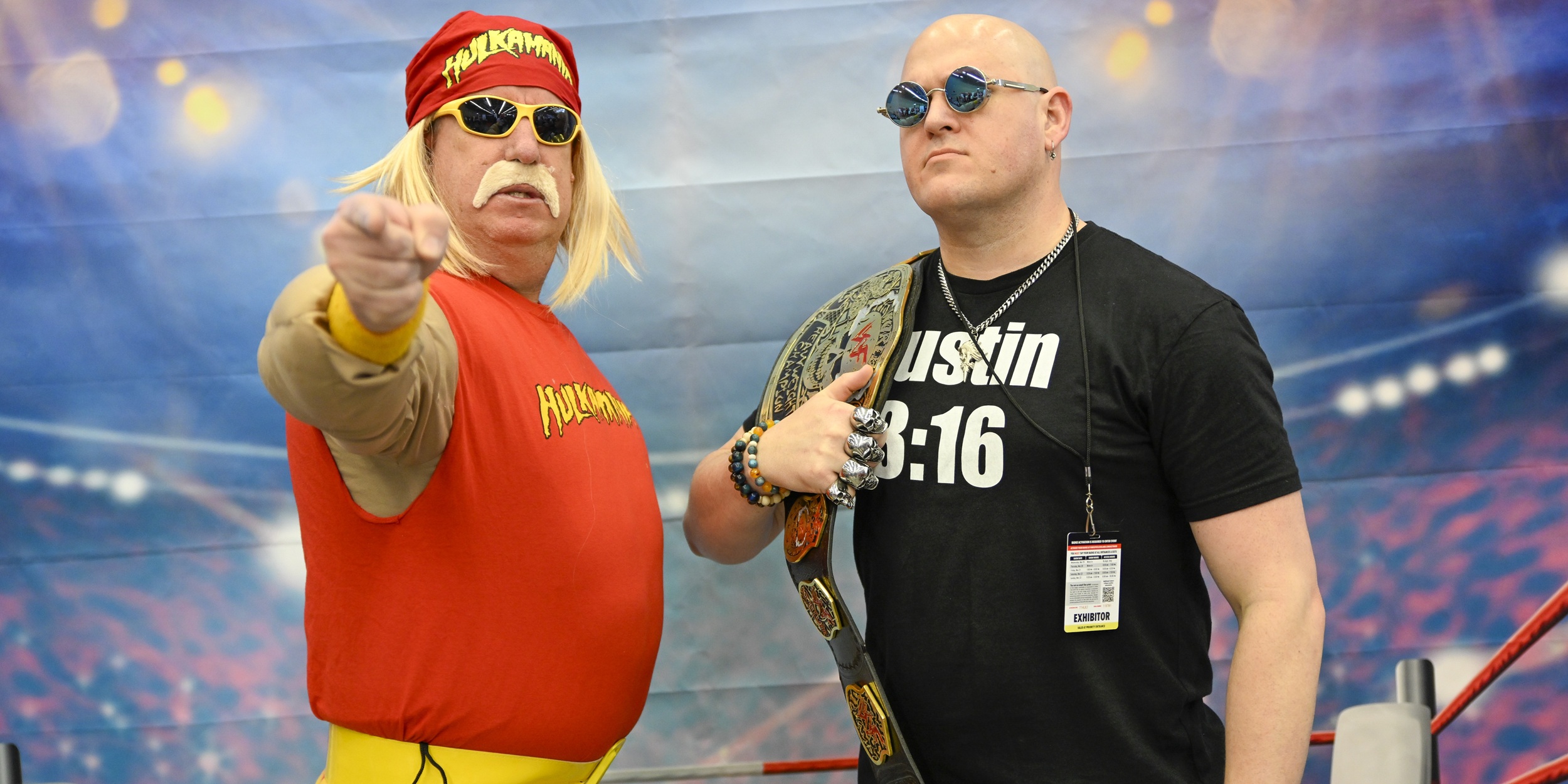 Two cosplayers pose confidently in front of a stadium-style backdrop. One wears a red sleeveless top and bandana with sunglasses, while the other stands beside him in a black shirt holding a championship-style belt.