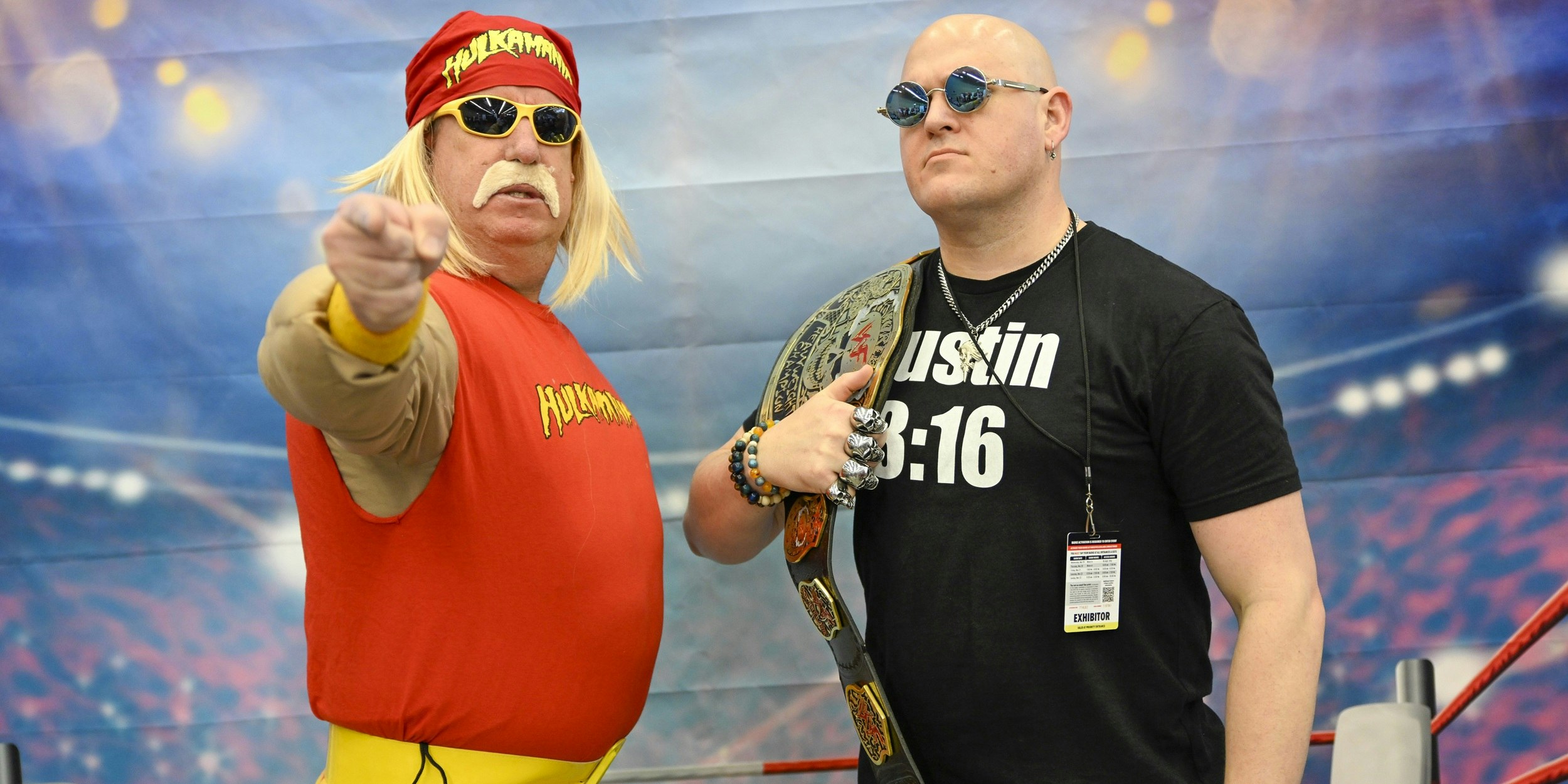 Two cosplayers pose confidently in front of a stadium-style backdrop. One wears a red sleeveless top and bandana with sunglasses, while the other stands beside him in a black shirt holding a championship-style belt.