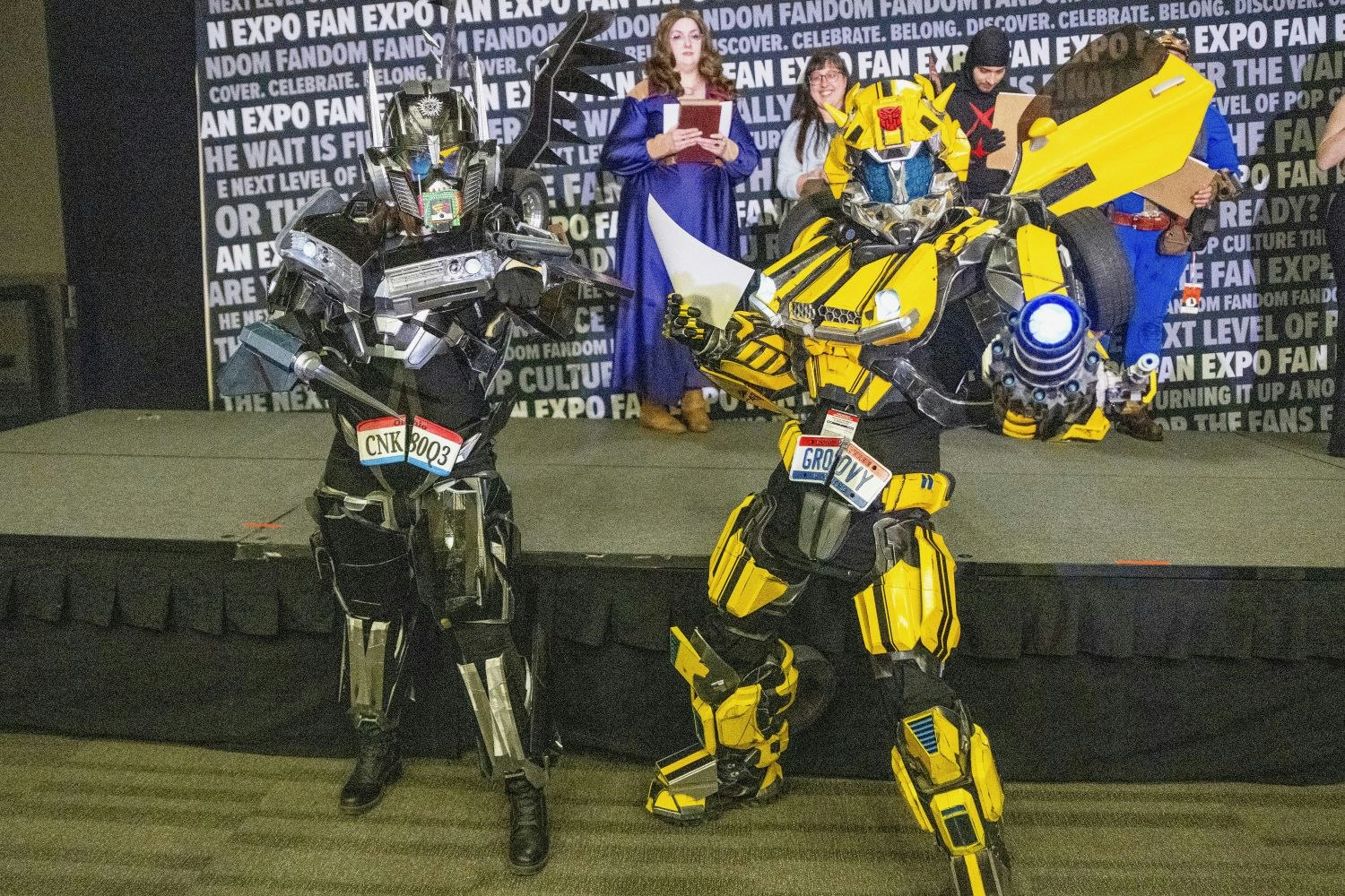 Two cosplayers dressed as Transformers-style robots pose on the convention floor—one in sleek black and silver armor, the other in yellow and black with a glowing cannon arm. Judges and other participants can be seen in the background.