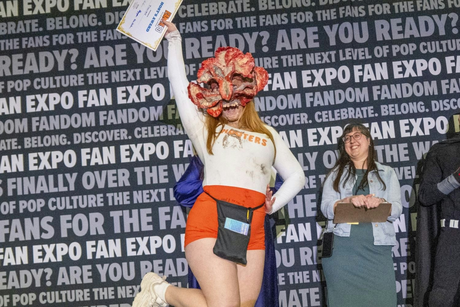 A cosplayer in a humorous crossover costume—a "Hooters" uniform with a grotesque Clicker mask from The Last of Us—excitedly raises a “Judge’s Award” certificate in the air while a judge in glasses smiles behind them.