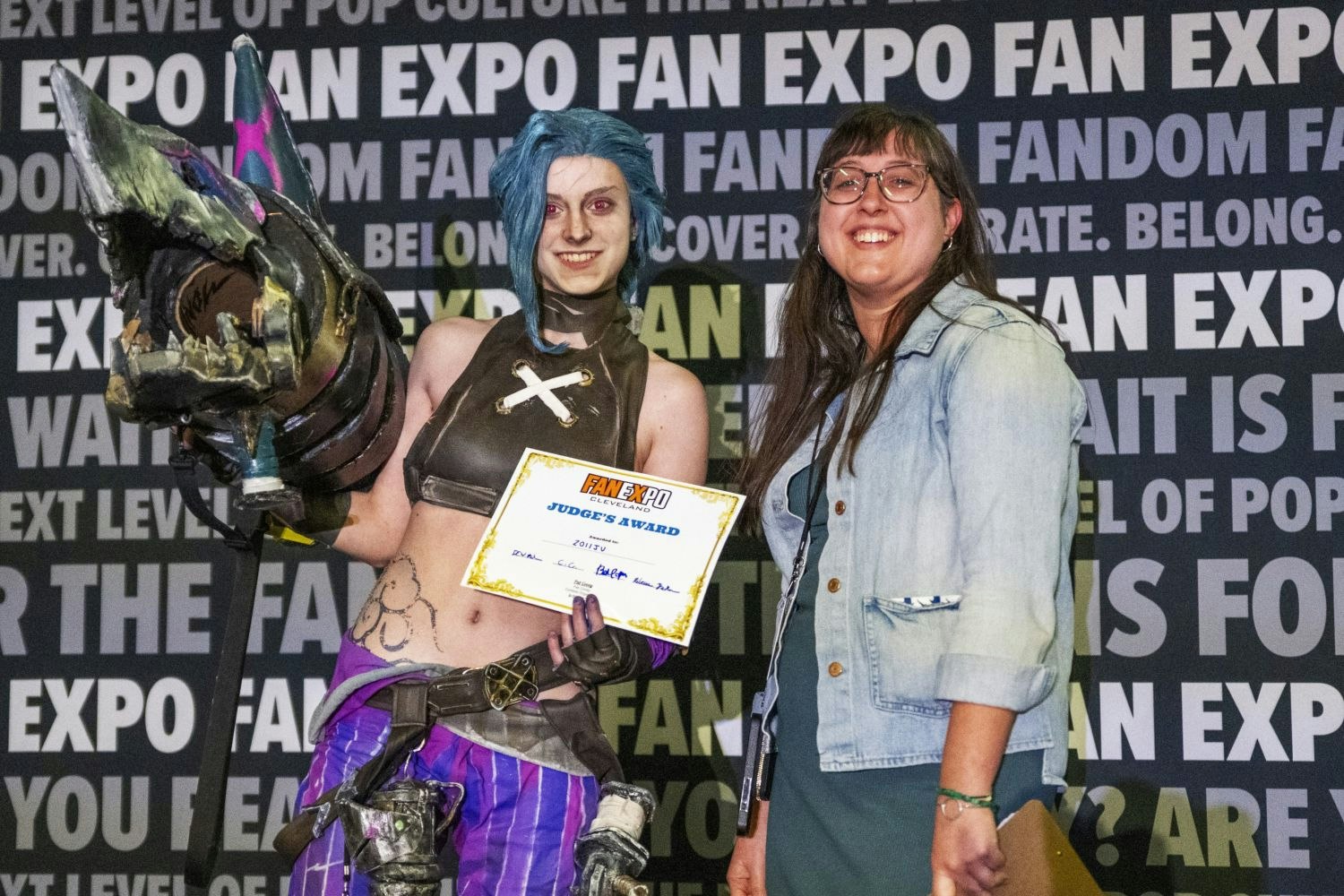 A cosplayer dressed as Jinx from Arcane poses with a massive shark-shaped weapon and a certificate reading "Judge’s Award" next to a smiling judge in glasses and a denim jacket.