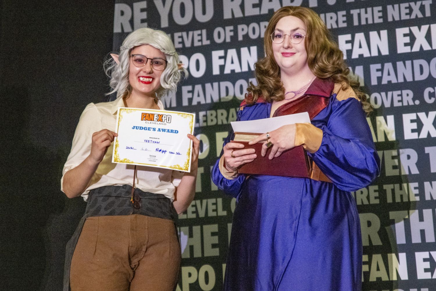 A cosplayer with pointed elf ears and white hair holds a “Judge’s Award” certificate while standing beside a judge dressed in a long, flowing purple and burgundy dress, holding a closed book.