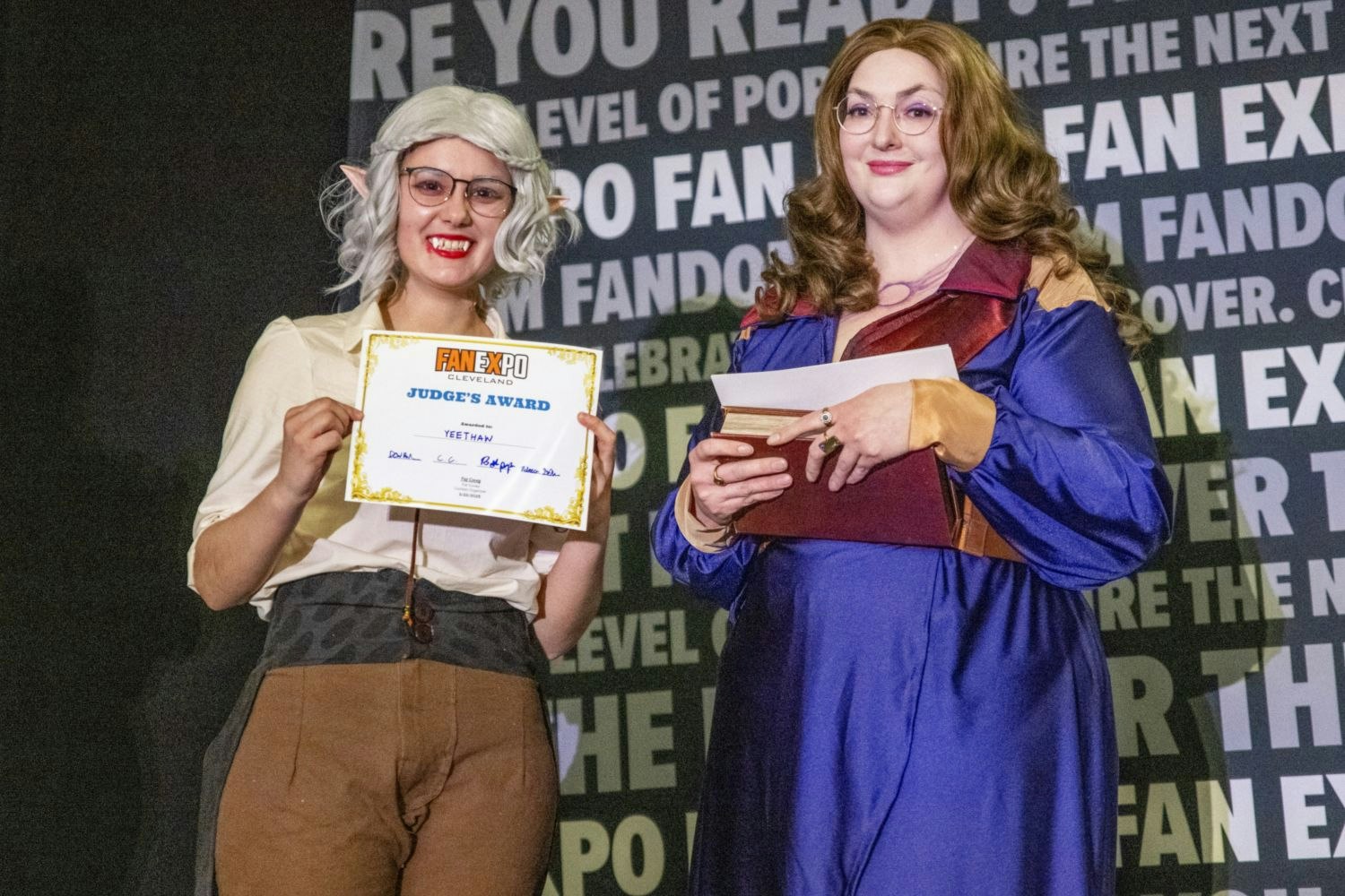 A cosplayer with pointed elf ears and white hair holds a “Judge’s Award” certificate while standing beside a judge dressed in a long, flowing purple and burgundy dress, holding a closed book.
