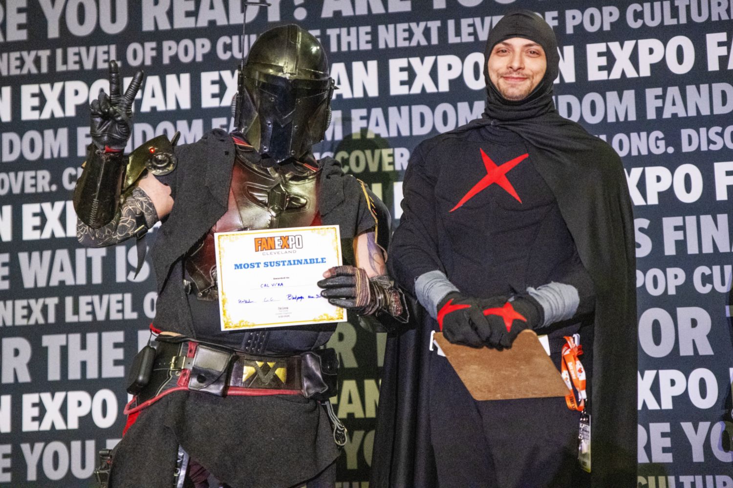 A cosplayer in a Mandalorian-inspired costume with black and red armor holds up a peace sign and a “Most Sustainable” certificate next to a judge in a black costume with a red “X” emblem.
