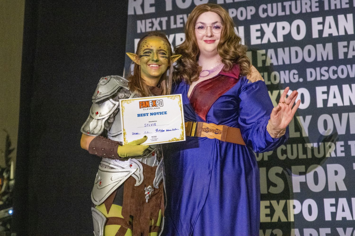 A cosplayer in green and silver elf-like armor with face paint holds a “Best Novice” certificate while smiling beside a judge in a long purple gown waving to the audience.