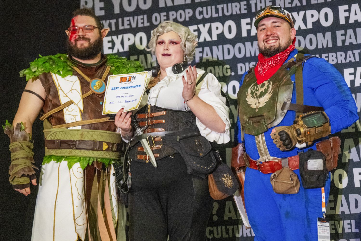 Three cosplayers stand together as a group, with the center cosplayer holding a “Best Journeyman” certificate. One is dressed in leafy green fantasy armor, one in a steampunk corset outfit, and one in a Fallout-inspired blue and red costume.