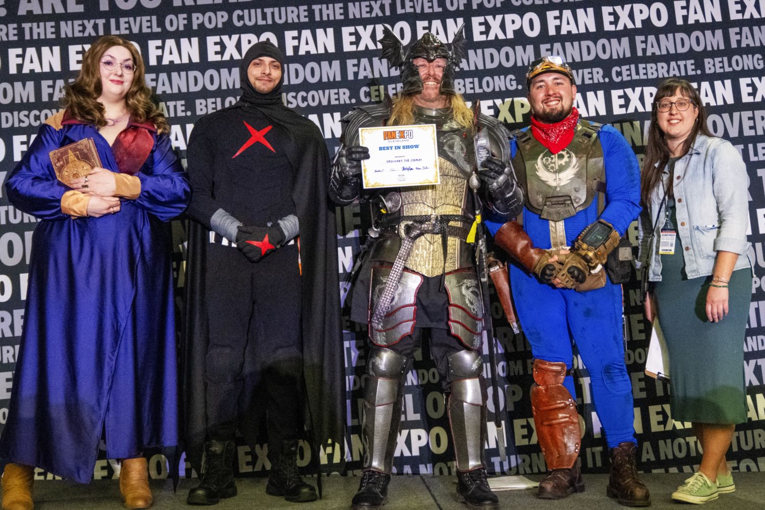 Five people stand in a row for a group photo. A heavily armored fantasy warrior in the center holds the “Best in Show” certificate. Judges stand around him, smiling proudly.