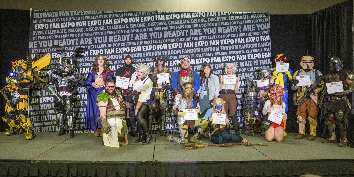 A large group of cosplayers pose together onstage at FAN EXPO Cleveland’s Cosplay Craftsmanship Cup. The backdrop behind them is covered with repeated FAN EXPO branding. The cosplayers showcase a wide mix of characters and genres, including armored sci-fi builds, fantasy heroes, video game characters, and pop-culture favorites. Many hold award certificates for categories like Best Novice, Best Master, and Best Group. Some wear full armor suits, others have elaborate props, wigs, or makeup, and a few kneel in the front row to display their costumes. The scene highlights the variety, creativity, and craftsmanship of the cosplay community at the show.