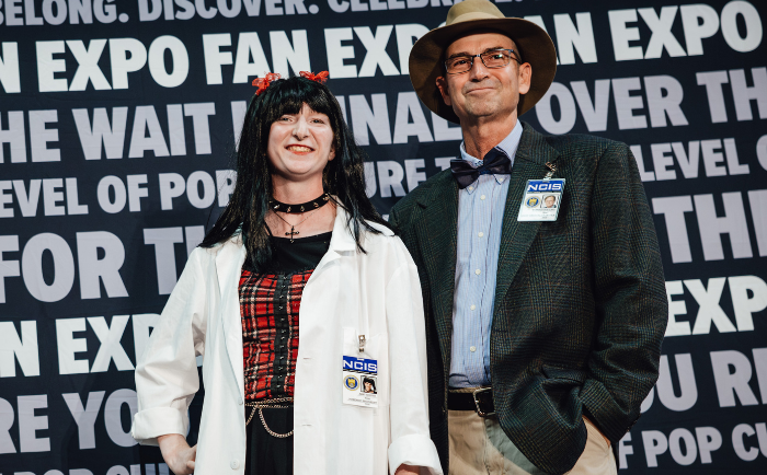 A pair stands against a "FAN EXPO" backdrop in cosplay attire. The woman on the left wears a white lab coat with an "NCIS" badge, a red plaid dress underneath, and a black choker necklace. Her black hair is accessorized with red bows. The man on the right sports a tweed jacket, a tan hat, glasses, and also displays an "NCIS" badge. Both exhibit proud smiles, showcasing their dedication to their character portrayals at the pop culture event.