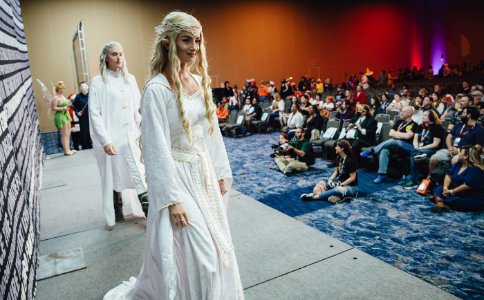 A woman and man in ethereal white gowns and intricate hairstyles stand on a stage. The woman in the forefront, adorned with a golden tiara, confidently looks forward with a gentle smile. The man, slightly behind, gazes into the distance. They're both in a large room with an audience of attendees seated and watching, some in their own colorful costumes. The atmosphere is that of a convention or cosplay event, with a stage backdrop to the left and ambient lighting illuminating the space.