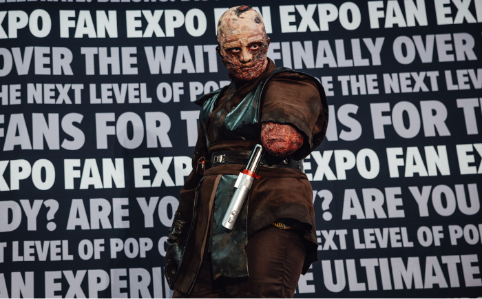 An individual in a detailed and gruesome cosplay stands against a "FAN EXPO" backdrop. The costume features a head covered in what appears to be bloody bandages with exposed areas showing scarred and burnt skin. They wear a dark, distressed outfit with a belt holding a metallic cylinder, possibly a prop weapon. The person's posture and makeup express dedication to portraying a horror or action character at a pop culture event.