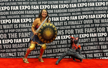 Two cosplayers posing on the Cosplay Red Carpet. On the left is a woman dressed as Wonder Woman, and on the right is a child dressed in a black Spider-Man costume.