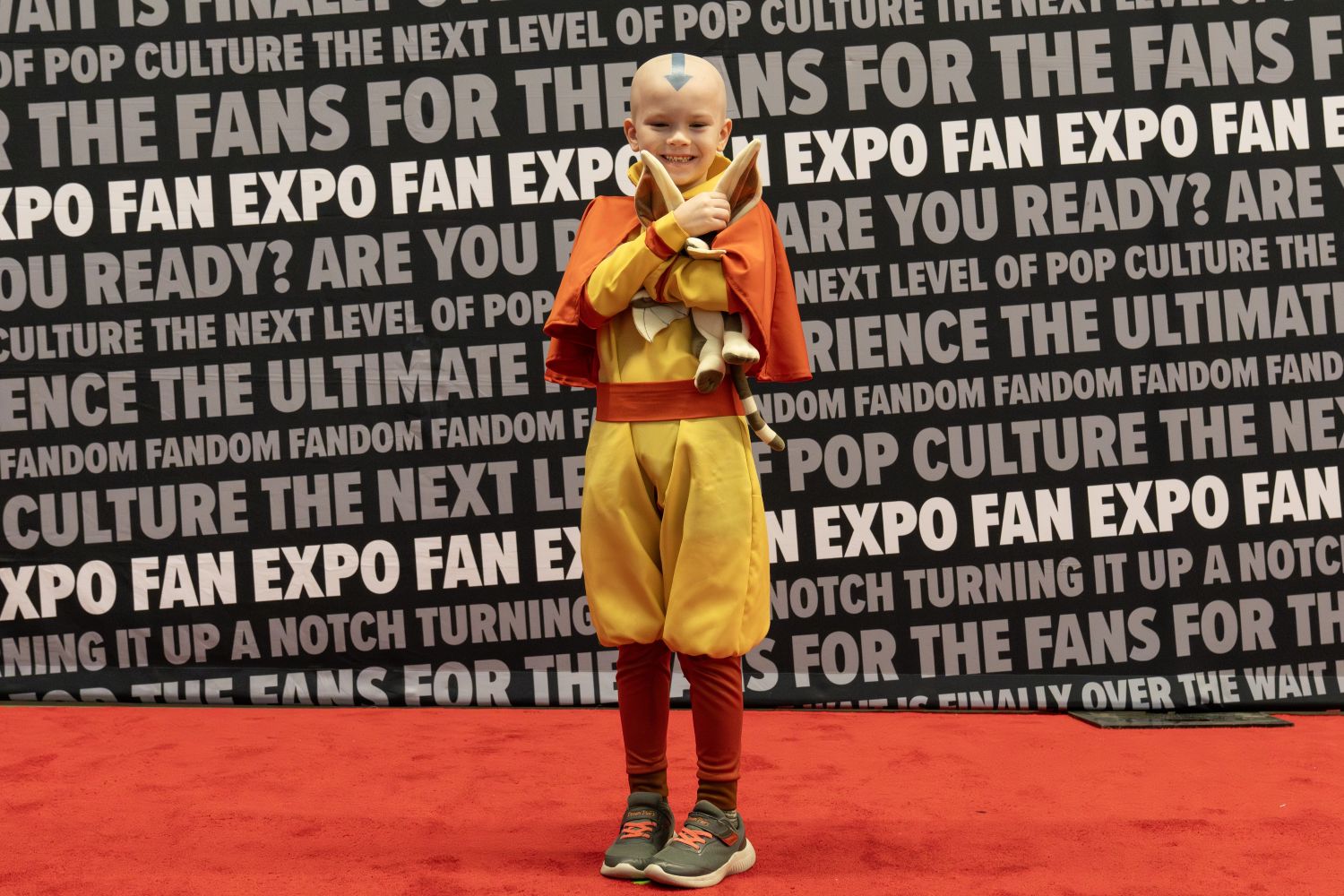 A child cosplayer beams as Aang from Avatar: The Last Airbender, complete with a painted blue arrow on their head and stuffed animal Momo tucked under their arm.