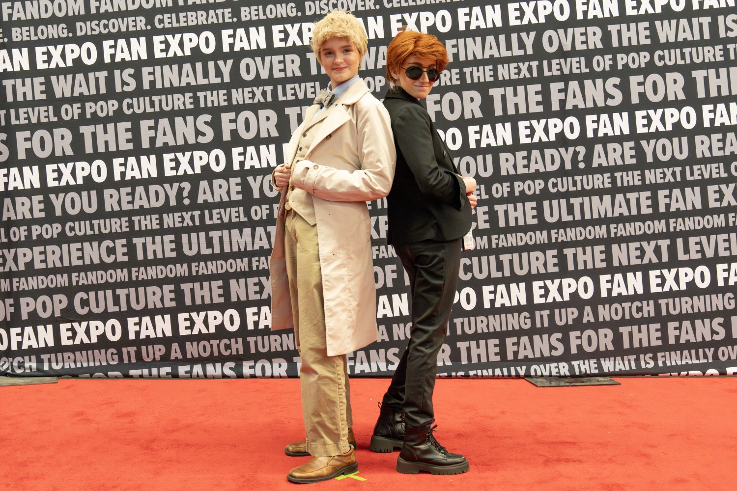 Two cosplayers pose back-to-back on the red carpet. One wears a light trench coat, vest, and slacks, resembling Aziraphale from Good Omens. The other, dressed in black with sunglasses and platform boots, channels Crowley from the same show.