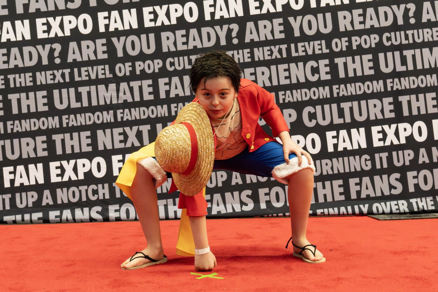 A young cosplayer strikes a dynamic pose as Monkey D. Luffy from One Piece, crouched low with a determined look, straw hat in hand, and wearing Luffy’s signature red jacket and blue shorts.