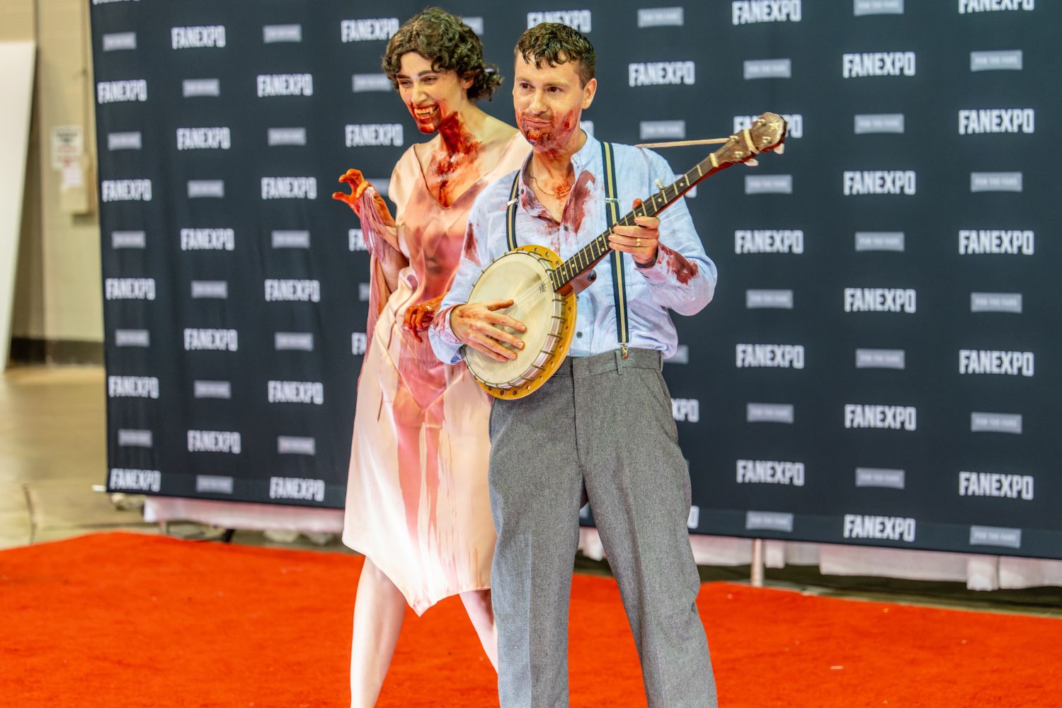 Two cosplayers in horror-themed attire stand on the red carpet. One wears a bloodied vintage dress with makeup resembling smeared blood, while the other, also blood-spattered, plays a banjo while wearing suspenders and trousers.