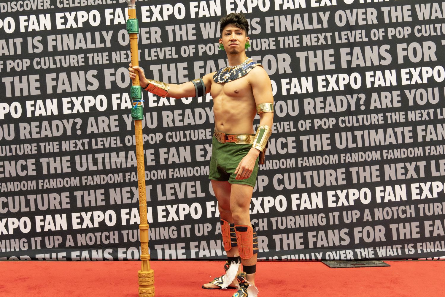 A cosplayer dressed as Namor from Black Panther: Wakanda Forever stands confidently on the red carpet holding a tall, ornate spear and wearing a regal costume with gold accents and green shorts.