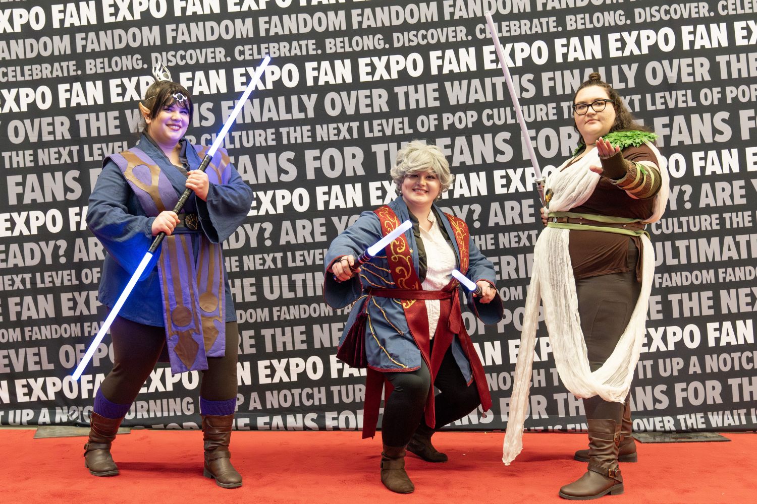 Three cosplayers in fantasy-inspired costumes pose with glowing lightsabers. Each is dressed as an original or mash-up Jedi character with detailed robes, armor, and accessories.