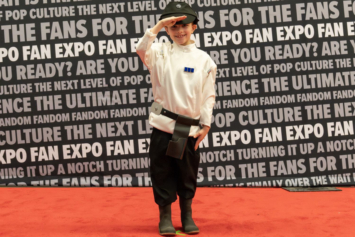A child cosplayer salutes on the red carpet dressed in an Imperial officer uniform from Star Wars, complete with a black hat and white tunic with rank insignia.