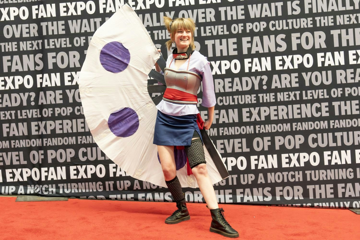 A cosplayer smiles in a powerful pose as Temari from Naruto, holding her iconic giant fan and wearing the character’s lavender outfit with fishnet leggings.