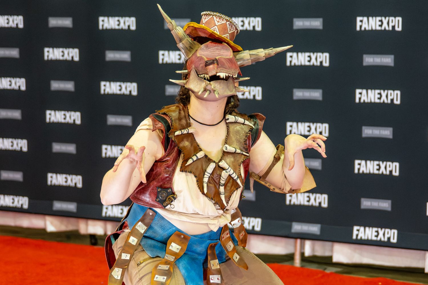 A cosplayer in an elaborate armored costume with horns, a jagged mask, and a tattered outfit crouches in character pose on the red carpet.