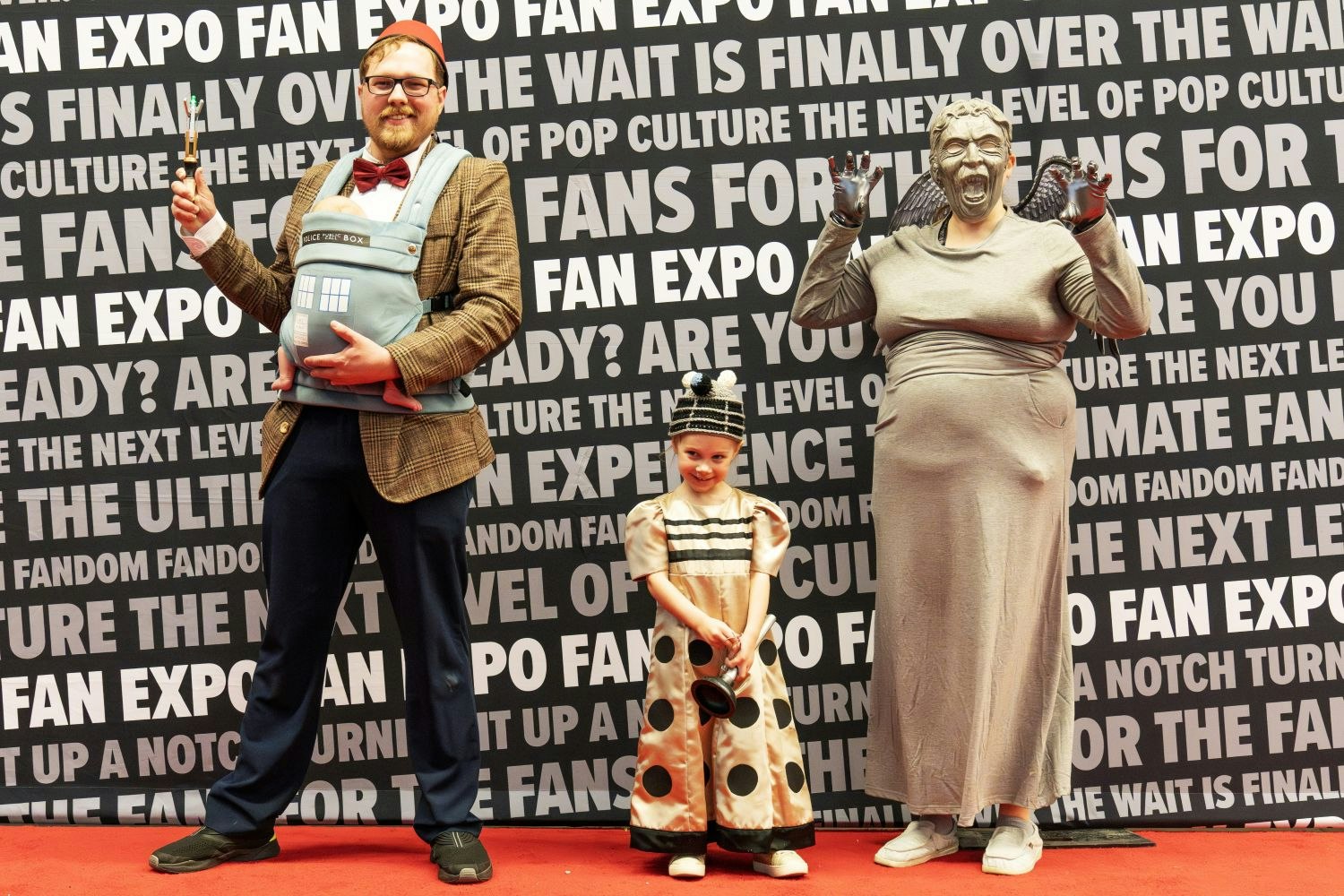 A family cosplay group strikes a pose: one dressed as the Eleventh Doctor from Doctor Who with a baby TARDIS in a carrier, a child in a Dalek costume, and another adult in a detailed Weeping Angel costume.