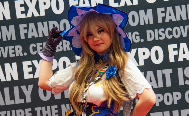 The image showcases a woman in an elaborate cosplay outfit at FAN EXPO. She is dressed as a character from a fantasy or anime series, featuring a large, decorative blue hat, long blonde wig, and a detailed costume with a white blouse, blue accents, and various ornaments. Her pose is confident and playful as she touches her hat and looks directly at the camera, fully embodying her character. The backdrop with text promoting the expo emphasizes the theme of fandom and pop culture, enhancing the setting of this cosplay showcase.