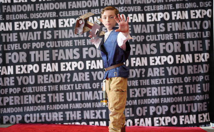 A young boy dressed in a blue vest and khaki pants stands confidently on a red carpet. He poses with one hand outstretched, showing a motion as if using the force, and holds a toy version of Han Solo's blaster in the other hand. The backdrop is filled with repeated phrases such as "FAN EXPO" and "FANDOM", indicating a pop culture event setting. The boy's expression is focused and serious, immersing himself in his character portrayal.