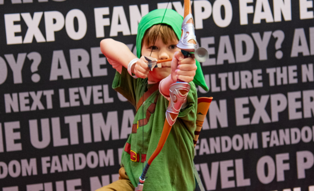 The image captures a young child dressed in a vibrant cosplay outfit at FAN EXPO. He is costumed as a well-known video game character, complete with a green tunic, hat, and a playful, oversized bow and arrow set. His stance is dynamic and focused, as he aims the bow at the camera, adding a lively and imaginative touch to the scene. The background is filled with text promoting the expo, reflecting the event's focus on pop culture and fandom, making it a perfect setting for attendees of all ages to showcase their creativity and enthusiasm.