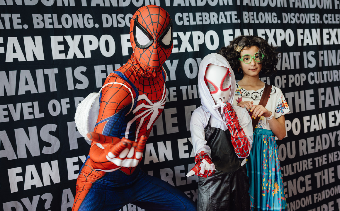 Two children and an adult pose in front of a "FAN EXPO" backdrop. The adult, dressed as Spider-Man, is in a dynamic crouching pose, showcasing the iconic Spider-Man hand gesture. One child, wearing a Spider-Gwen costume, stands next to him with a white mask covering the face. The other child, to the right, sports green round glasses and is dressed in a colorful dress adorned with illustrations of cacti, while confidently holding a pair of red heart-shaped sunglasses. All three are clearly enjoying their time at a pop culture event.