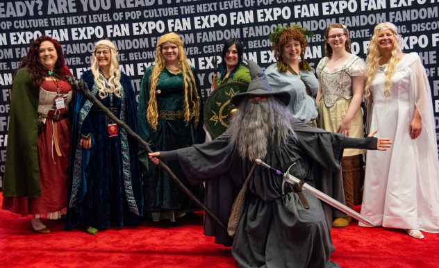 The image features a group of individuals at FAN EXPO, dressed in elaborate fantasy-themed costumes, reminiscent of characters from epic tales and legends. In the foreground, a person in a wizard costume dramatically points a staff forward, wearing a long gray robe and a pointed hat. Behind the wizard, six others are arrayed in a variety of richly detailed costumes, including regal dresses and cloaks, each adorned with thematic accessories like crowns, brooches, and embroidered patterns. Their expressions range from joyous to majestic, enhancing the fantastical atmosphere of the cosplay event. The group stands on a red carpet, with the event's logo and themes of pop culture displayed in the background, highlighting the vibrant and creative spirit of the convention.