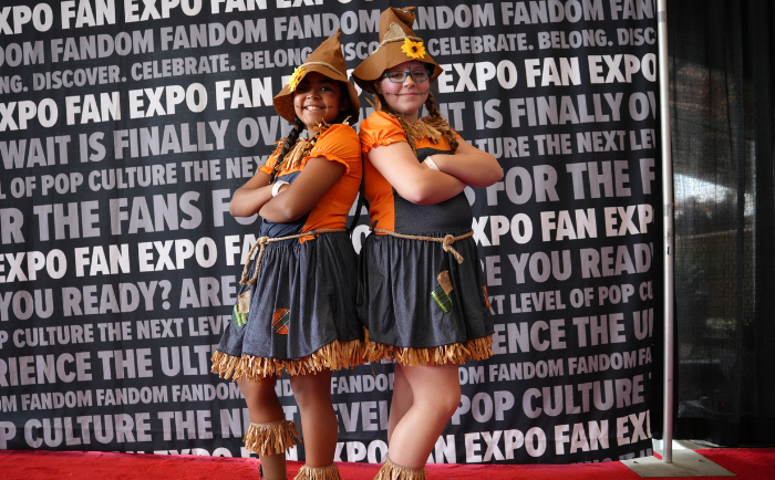 Two young girls stand confidently in front of a "FAN EXPO" backdrop. Both are dressed in matching outfits that consist of bright orange shirts, grey skirts with fringes at the bottom, and accessorized with brown belts holding small tools. They wear matching brown hats with orange decorations. Both girls have their arms crossed, exuding a playful and spirited demeanor, as they enjoy their time at a pop culture event.