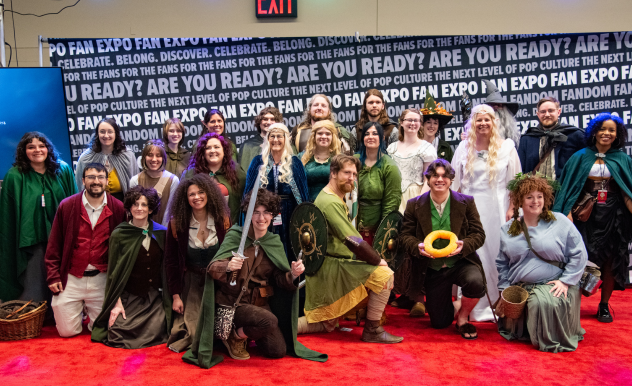 The image showcases a large group of people at FAN EXPO, gathered for a group photo, all dressed in a diverse array of fantasy and medieval-themed costumes. The participants are smiling and posing in various stances, from kneeling to standing, each displaying unique elements of their intricate outfits, such as cloaks, robes, and fantastical accessories like staffs and shields. The group's enthusiastic expressions and the detailed, colorful costumes create a lively and immersive atmosphere typical of a cosplay event. The background features vibrant promotional text about the fan culture, enhancing the festive and community-centric vibe of the occasion.
