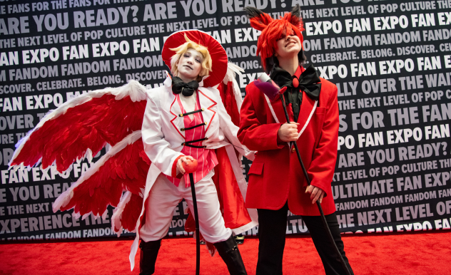 The image features two individuals in vibrant and dramatic cosplay at FAN EXPO. The person on the left is dressed in a white suit with detailed red trim, complemented by a large, feathered white wing and a matching cap, exuding an angelic theme. The person on the right wears a striking red suit with a high-collared coat and also holds a microphone, their bright orange hair styled upward dramatically. Both cosplayers strike playful poses on a red carpet, their costumes vivid against the backdrop adorned with FAN EXPO logos and text related to fan culture, enhancing the celebratory and creative atmosphere of the event.