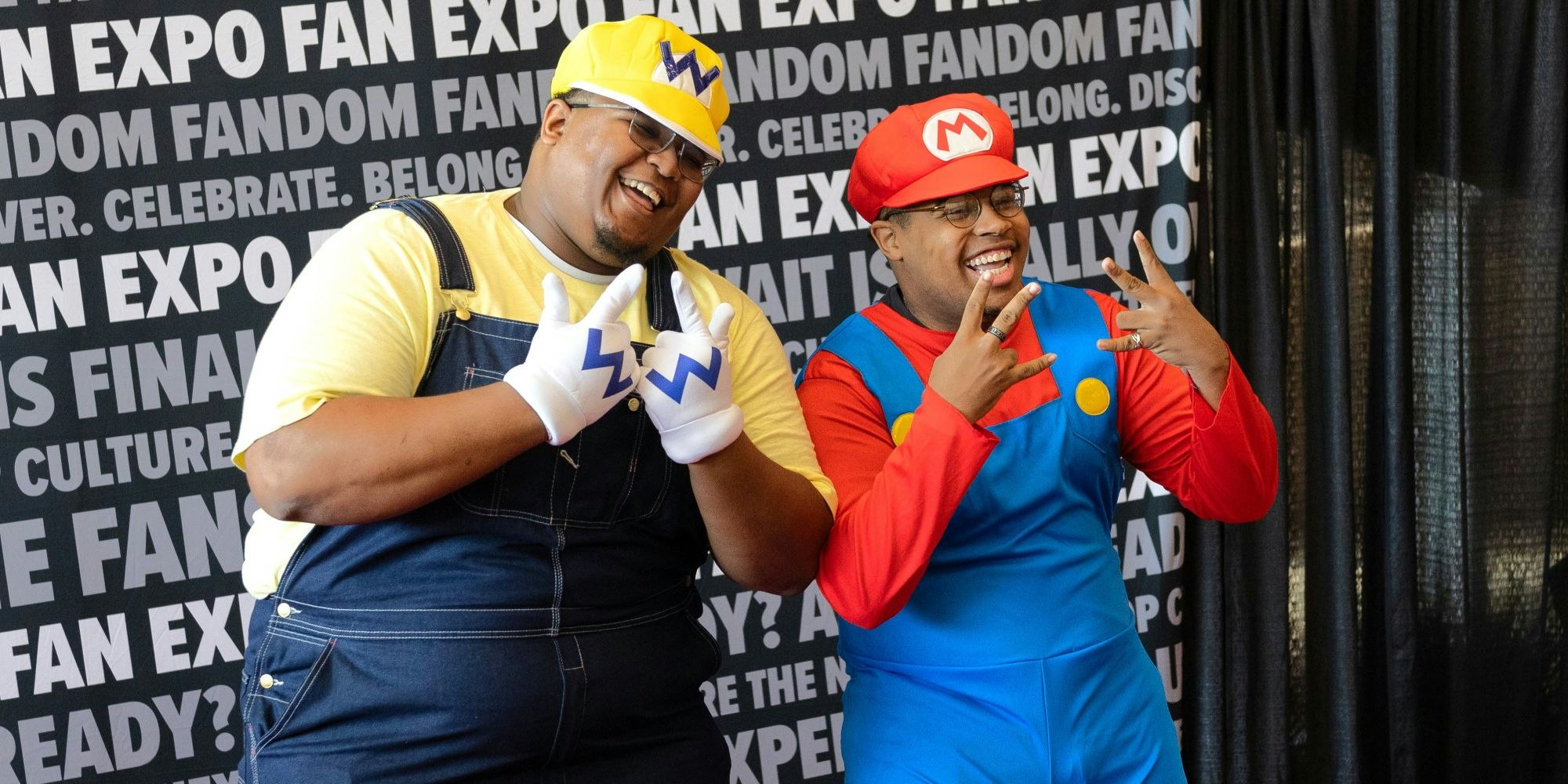 Two fans dressed as Mario and Wario pose together at Dallas FAN FESTIVAL. They smile and throw playful hand signs while wearing colorful overalls, hats, and gloves in front of a FAN EXPO backdrop.