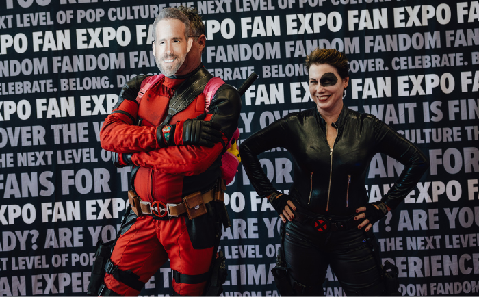 Two individuals pose confidently in front of a backdrop plastered with "FAN EXPO" and other fandom-related phrases. The person on the left is dressed as Deadpool, in a red and black tactical suit with arms crossed, while the person on the right is outfitted as Domino, sporting a sleek black leather costume with a prominent X-symbol on the belt. Both display joyful expressions, immersing themselves in their characters.