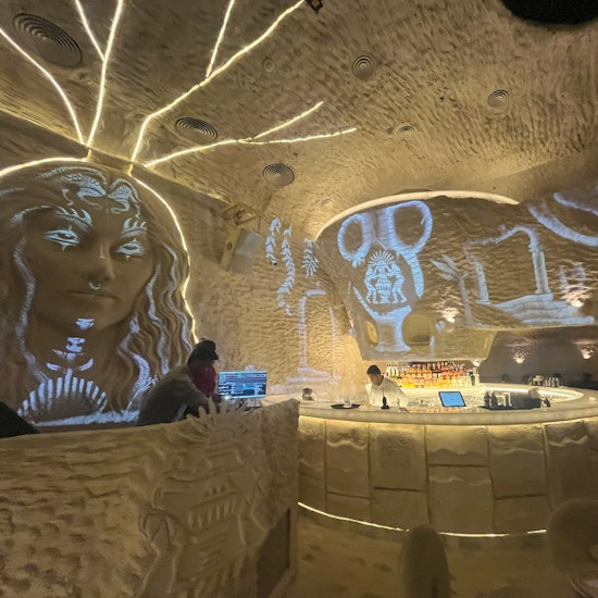 Futuristic cave-style bar interior with illuminated wall art projections, including a large face and symbolic designs, behind a circular counter where bartenders serve drinks