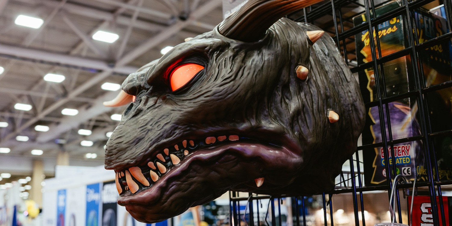 A large, monstrous creature prop with sharp teeth and glowing eyes hangs behind a display booth. The detailed sculpting and textured surface create a menacing appearance as part of the Cursed Objects exhibit.