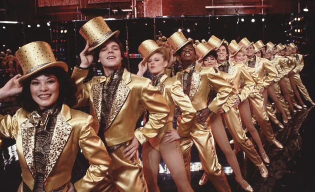 Standing in a 15 person kick-line, the chorus line wear all gold outfits and hats as they extend their legs during a dance number