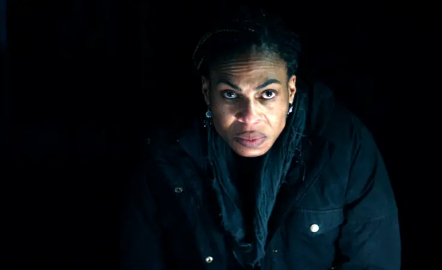 A woman wearing a black winter coat and grey scarf stares menacingly at the camera.