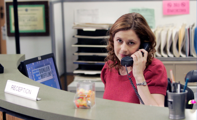 Pam sits behind the reception desk, she looks toward the camera as she answers the phone.