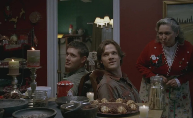 Sam and Dean sit tied up at a table decked out for the Holidays - A woman in a Christmas sweater leans in to speak to them.