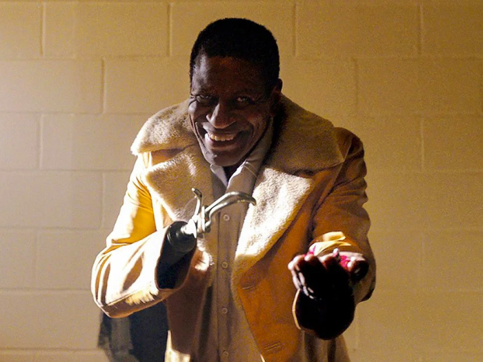 A man with a beige fluffy jacket and a metal hook smiles ominously at the camera