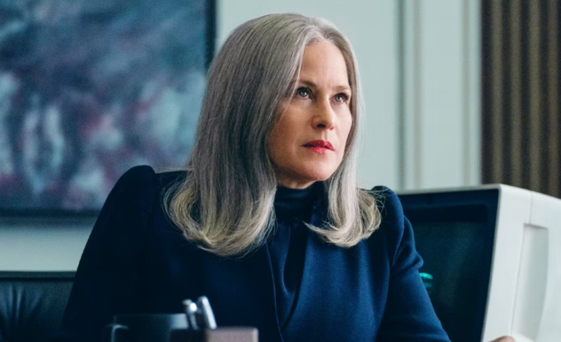 Harmony sits at her desk, her clothing, an the items around her are all varying shades of blue. As she looks to a person off screen, she is serious.