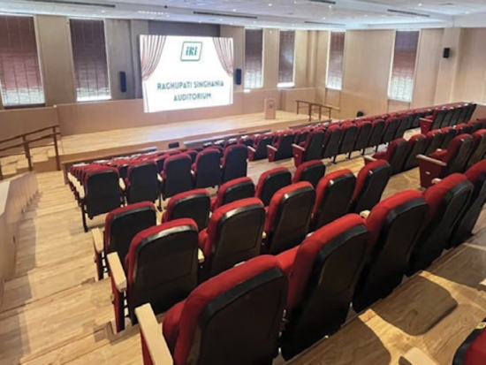 Conference hall with red seating, viewed from the back, facing a presentation screen at the front