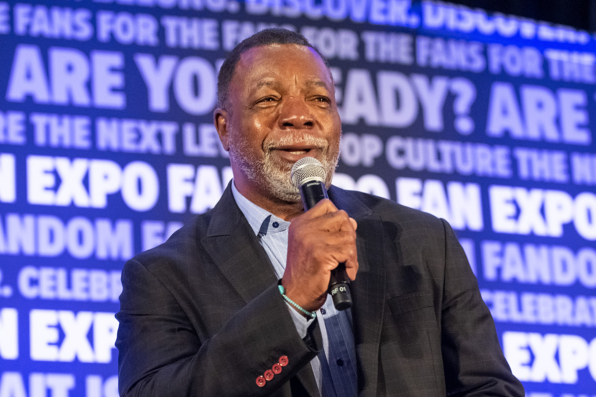 A head shot of Carl Weathers holding a microphone. He is smiling as he answers a question. His has buzzed short black hair and a short white beard. He is wearing a black suit jacket with a light blue button up. On his wrist is a teal bracelet with a circular middle charm. In the background, is the FAN EXPO background that repeats a variety of phrases.